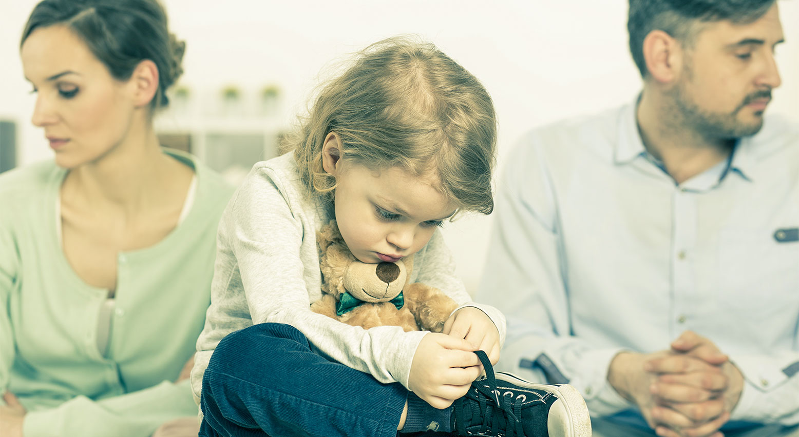 Foto einer Familie: Ein Kind hält einen Teddy im Arm, dahinter sitzen Mutter und Vater und blicken in entgegengesetzte Richtungen. Alle schauen traurig.