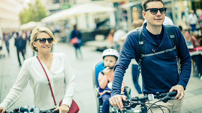 Elternpaar mit Kleinkind schiebt Fahrräder durch Fußgängerzone.